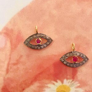 Puede incluir: Un par de pendientes de oro de ojo turco con una piedra de rubí roja en el centro de cada ojo. Los pendientes están rodeados de pequeños diamantes.