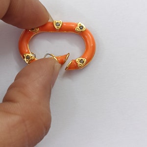 Op de afbeelding: Ovale sluiting van oranje emaille en goudkleurig metaal met kleine diamant-achtige accenten.