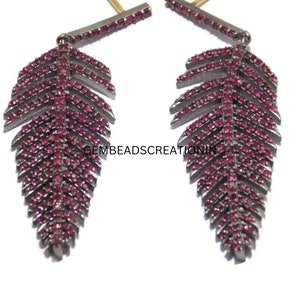 Puede incluir: Un par de pendientes colgantes con un diseño de hoja en metal oscuro incrustado con pequeñas piedras rojas. Cada pendiente cuelga de una fina barra horizontal también decorada con piedras rojas. El texto GEMBEADSCREATIONIN es visible.