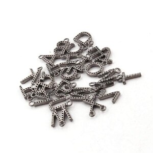 May include: A pile of small, silver-toned alphabet charms. Each charm represents a different letter, with a small loop for attaching to jewellery. The letters are dotted with small holes, and some have small, clear stones.
