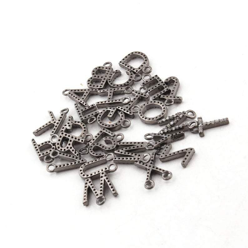 May include: A pile of small, silver-toned alphabet charms. Each charm represents a different letter, with a small loop for attaching to jewellery. The letters are dotted with small holes, and some have small, clear stones.