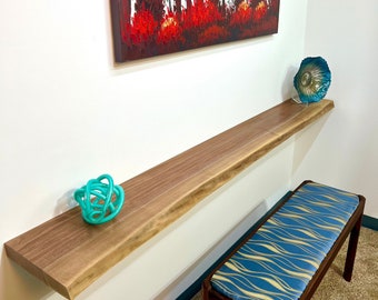 May include: A wooden shelf with a live edge, mounted on a white wall. The shelf is decorated with a turquoise glass orb and a blue and white seashell. A wooden bench with a blue and gold patterned cushion sits in the foreground.