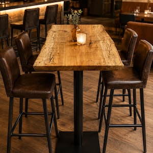 May include: A rustic wooden bar table with a live edge, supported by a black metal base. Four brown leather bar stools with black metal legs surround the table. A small candle and a vase with greenery sit on the table. The bar is in the background.