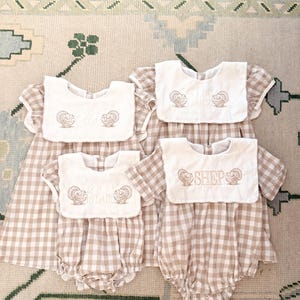Può includere: Quattro completi per neonati con motivo a quadretti beige e bianco. Ogni completo include un bavaglino abbinato con testo ricamato e disegni di tacchini. I bavaglini hanno i nomi "Ella", "Lainie", "Blair" e "Shep".