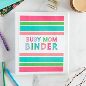 Pode incluir: Uma pasta branca com uma capa colorida que diz "Busy Mom Binder". A capa tem listras rosa, verdes e azuis.