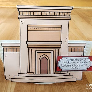 May include: A paper craft of a building with a tan facade, brown trim, and a central arched doorway. A sign with text from Psalm 127:1 is in front. The building is on a white plate, set on a wooden surface.