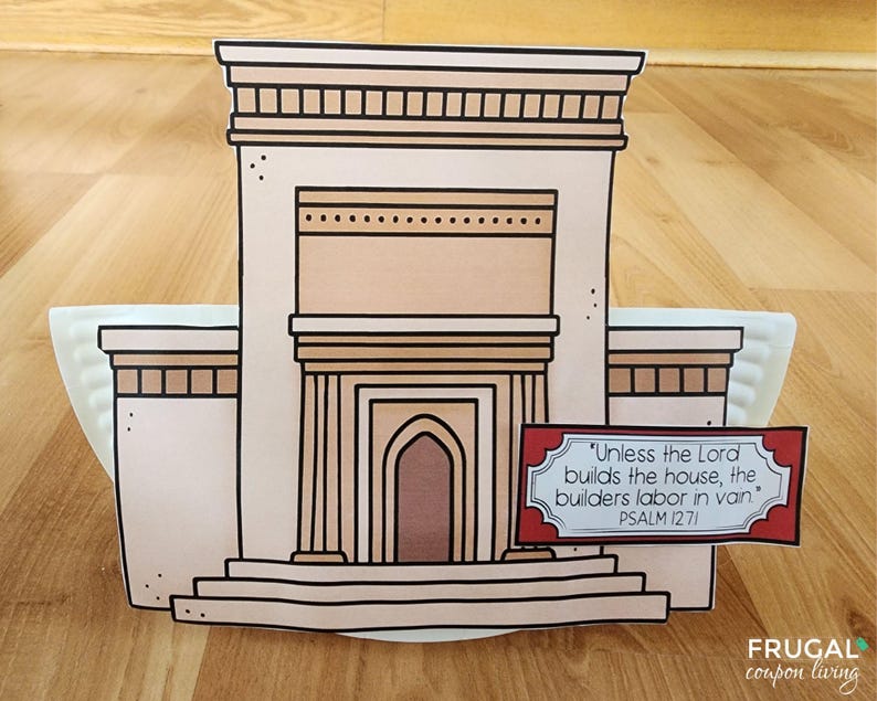 May include: A paper craft of a building with a tan facade, brown trim, and a central arched doorway. A sign with text from Psalm 127:1 is in front. The building is on a white plate, set on a wooden surface.