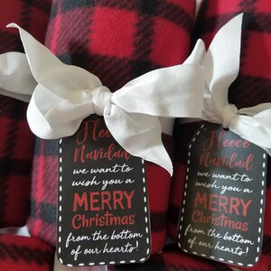 May include: Rolled red and black plaid fleece blankets tied with white ribbon bows. Each blanket has a black tag with white dotted edges and the text "Fleece Navidad, Merry Christmas from the bottom of our hearts!"