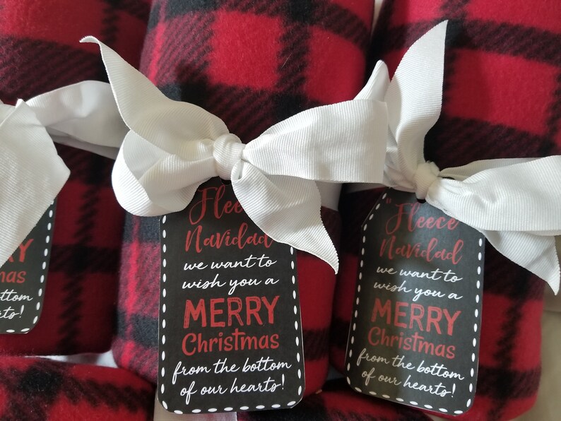 May include: Rolled red and black plaid fleece blankets tied with white ribbon bows. Each blanket has a black tag with white dotted edges and the text "Fleece Navidad, Merry Christmas from the bottom of our hearts!"