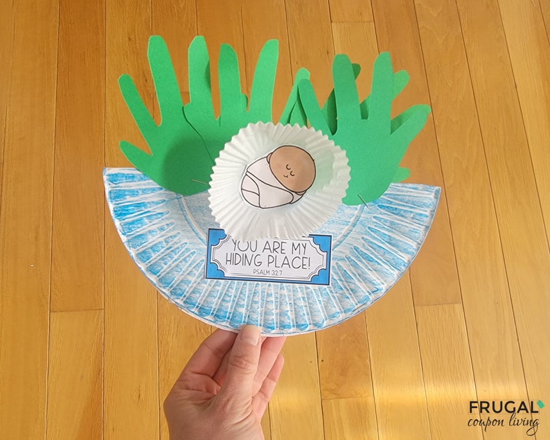 Pu&ograve; includere: Un piatto di carta blu con un pirottino da cupcake bianco al centro. Il pirottino da cupcake contiene un disegno di un bambino all'interno. Due impronte di mani di carta verde sono sopra il pirottino da cupcake. Il piatto di carta ha un bordo a strisce blu e bianco. Un cartello blu e bianco con il testo "You are my hiding place! Psalm 32:7" &egrave; sotto il pirottino da cupcake.