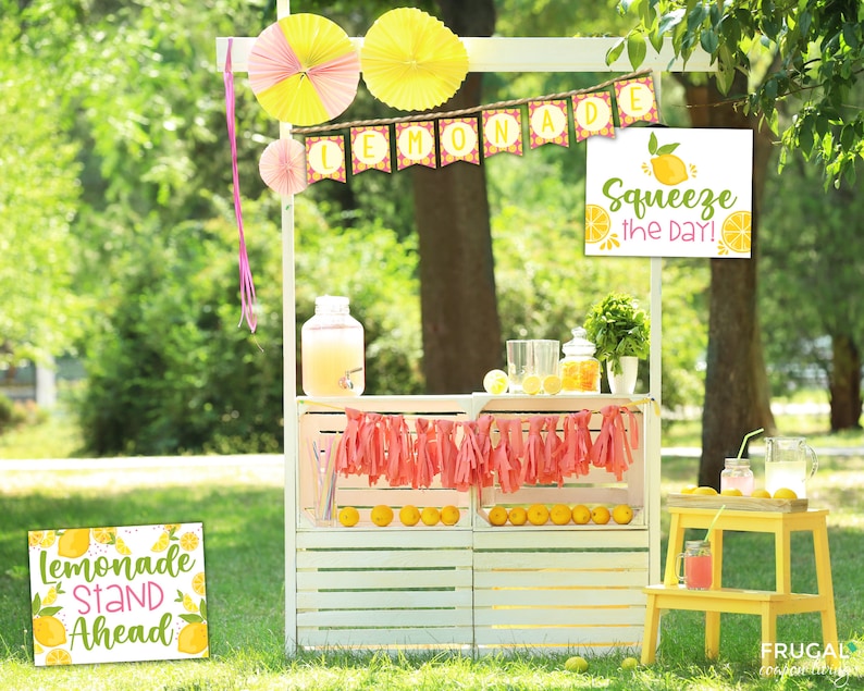 May include: A white lemonade stand decorated with yellow and pink paper fans and a banner that says "Lemonade." A sign reads "Squeeze the Day!" and another says "Lemonade Stand Ahead." Lemons and lemonade are visible.
