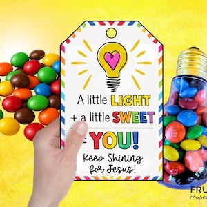 May include: A colorful gift tag with a lightbulb design and the text "A little LIGHT + a little SWEET = YOU! Keep Shining for Jesus!". A hand holds the tag next to a pile of candies and a lightbulb-shaped container filled with candies.