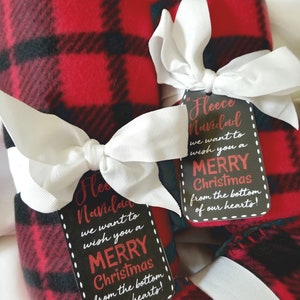 May include: Red and black plaid fleece blankets tied with white bows and gift tags. The tags read "Fleece Navidad, we want to wish you a Merry Christmas from the bottom of our hearts!" A festive holiday gift.