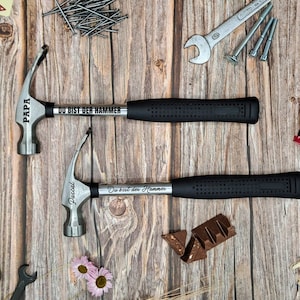 Könnte beinhalten: Zwei silberne Krallenhämmer mit schwarzen Griffen auf einem Holzgrund. Die Hämmer sind mit den Worten "Papa" und "Du bist der Hammer" sowie "Pascal" und "Du bist der Hammer" graviert.