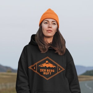 Könnte beinhalten: Ein schwarzer Hoodie mit einer orangenen Berggrafik und dem Text "DER BERG RUFT" in einer Raute. Das Model trägt den Hoodie mit schwarzen Leggings und braunen Stiefeln.