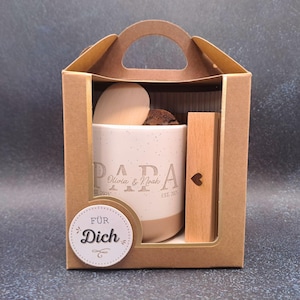 May include: A gift set in a brown cardboard box. Inside, a beige mug with the word "PAPA" and names, a wooden heart, and a wooden stick with a heart cutout. A sticker says "F&Uuml;R Dich".