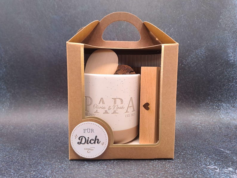 May include: A gift set in a brown cardboard box. Inside, a beige mug with the word "PAPA" and names, a wooden heart, and a wooden stick with a heart cutout. A sticker says "F&Uuml;R Dich".