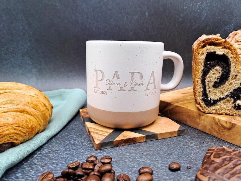 May include: White ceramic mug with the word "PAPA" and the names "Olivia & Noah" engraved on it. The mug has a tan base and is on a wooden coaster. A croissant, bread, and coffee beans are also visible.