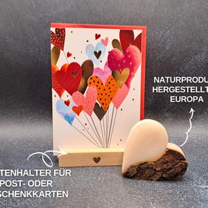 May include: A greeting card with a red border and a colourful heart design, displayed in a wooden card holder. A wooden heart-shaped decoration with bark is next to the card. Text on the card holder reads "Kartenhalter f&uuml;r Post- oder Geschenkkarten".
