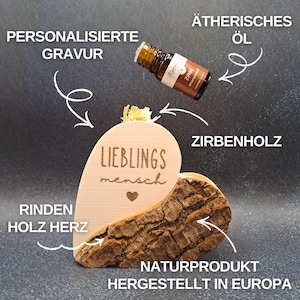 Peut inclure: Décoration en bois en forme de cœur avec l'inscription "LIEBLINGS mensch" gravée. Elle est faite de bois naturel et d'écorce, et comprend une petite bouteille d'huile essentielle. Le texte "NATURPRODUKT HERGESTELLT IN EUROPA" est également visible.