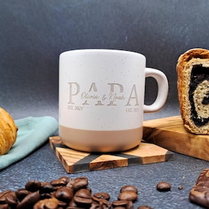 May include: A white and beige ceramic mug with the word "PAPA" engraved on the front, along with the names "Olivia & Noah" and the dates "EST. 2021" and "EST. 2025". The mug is on a wooden coaster, with coffee beans, a croissant, and bread in the background.