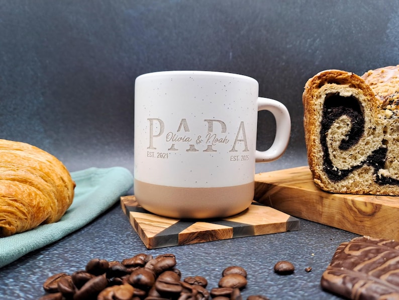 May include: A white and beige ceramic mug with the word "PAPA" engraved on the front, along with the names "Olivia & Noah" and the dates "EST. 2021" and "EST. 2025". The mug is on a wooden coaster, with coffee beans, a croissant, and bread in the background.