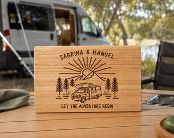 Könnte beinhalten: Ein rechteckiges Schneidebrett aus hellem Holz mit einer Lasergravur, die ein Wohnmobil, Tannenbäume und eine über Bergen aufgehende Sonne zeigt. Der Text lautet oben SABRINA & MANUEL und unten LET THE ADVENTURE BEGIN.