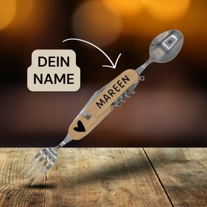 Könnte beinhalten: Ein Besteck mit Holzgriff, an dem sich an den Enden eine Gabel und ein Löffel befinden. Der Griff ist mit dem Wort "MAREEN" graviert. Ein kleines Herz ist in den Griff graviert. Das Besteck liegt auf einer Holzoberfläche.