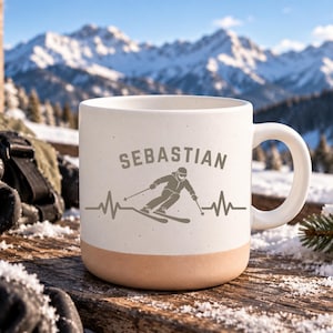 Op de afbeelding: Een witte keramische mok met een gespikkelde afwerking en een beige basis. De voorzijde toont een grijze afbeelding van een skiër verwerkt in een hartslaglijn, met de naam SEBASTIAN in grijze blokletters boven de ski-illustratie.