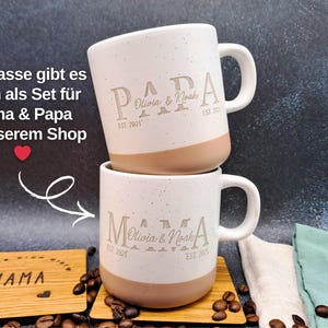May include: Two white ceramic mugs with a speckled finish and tan base, stacked. The mugs are engraved with the word "PAPA", a name, and "EST. 2021". The mugs are on a wooden coaster. The text on the image says "Die Tasse gibt es auch als Set f&uuml;r Mama & Papa in unserem Shop".