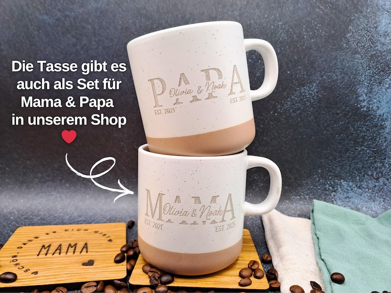 May include: Two white ceramic mugs with a speckled finish and tan base, stacked. The mugs are engraved with the word "PAPA", a name, and "EST. 2021". The mugs are on a wooden coaster. The text on the image says "Die Tasse gibt es auch als Set f&uuml;r Mama & Papa in unserem Shop".