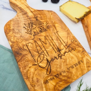 May include: Wooden chopping board with a handle, featuring the engraved word "FAMILIE" and "Gartner" with a house illustration. Cheese, blackberries, and a teal napkin surround the board. Coordinates are also engraved.