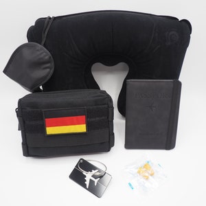 May include: A black travel set including a neck pillow, eye mask, passport holder, pouch with a German flag patch, earplugs, and a silver airplane luggage tag. The items are arranged on a white surface.