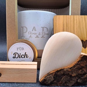 May include: A gift set including a white speckled ceramic mug engraved with "PAPA Olivia & Noah EST. 2021", a wooden heart, a wooden coaster with a heart cutout, and a small wooden block with a heart. A round sticker says "F&Uuml;R Dich".