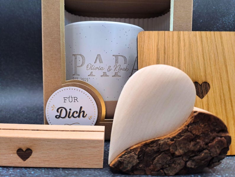 May include: A gift set including a white speckled ceramic mug engraved with "PAPA Olivia & Noah EST. 2021", a wooden heart, a wooden coaster with a heart cutout, and a small wooden block with a heart. A round sticker says "F&Uuml;R Dich".