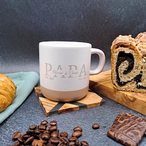 May include: A white ceramic mug with the word "PAPA" and the names "Olivia & Noah" printed on it. The mug sits on a wooden coaster, next to a croissant, coffee beans, and a loaf of bread.