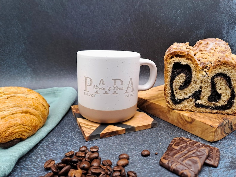 May include: A white ceramic mug with the word "PAPA" and the names "Olivia & Noah" printed on it. The mug sits on a wooden coaster, next to a croissant, coffee beans, and a loaf of bread.