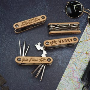 Könnte beinhalten: Mehrere Holz-Multitools mit Fahrradmotiven und Text, darunter "Gute Fahrt Wir Lieben Dich" und "HARRY". Die Werkzeuge sind geöffnet und zeigen verschiedene Werkzeuge. Sonnenbrille, Karte und Kompass sind ebenfalls sichtbar.