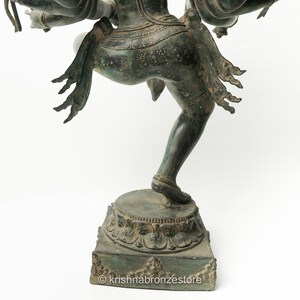 May include: A bronze statue of a Hindu deity with multiple arms, standing on a square base with intricate carvings. The statue is in a dancing pose, with one leg raised and the other bent. The deity is wearing a long skirt and a crown.