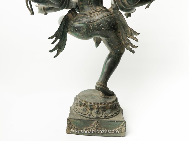 May include: A bronze statue of a Hindu deity with multiple arms, standing on a square base with intricate carvings. The statue is in a dancing pose, with one leg raised and the other bent. The deity is wearing a long skirt and a crown.