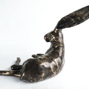 K&ouml;nnte beinhalten: Eine Bronzeskulptur eines liegenden Hasen mit seitlich gedrehtem Kopf und hoch erhobenen langen Ohren. Die Skulptur hat eine strukturierte Oberfl&auml;che und eine dunkelbraune Farbe.