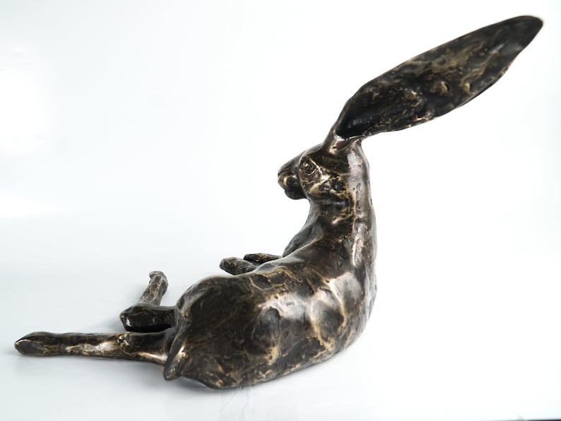 K&ouml;nnte beinhalten: Eine Bronzeskulptur eines liegenden Hasen mit seitlich gedrehtem Kopf und hoch erhobenen langen Ohren. Die Skulptur hat eine strukturierte Oberfl&auml;che und eine dunkelbraune Farbe.