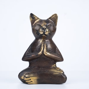 May include: A bronze cat figurine in a seated meditation pose with its paws folded in front of its chest. The cat has a serene expression with closed eyes.