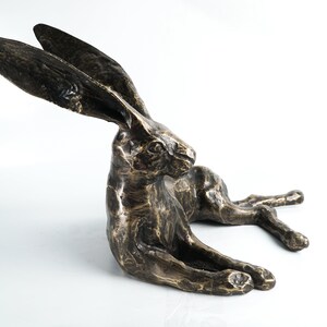 K&ouml;nnte beinhalten: Eine Bronzeskulptur eines liegenden Hasen mit seitlich gedrehtem Kopf. Der Hase hat lange Ohren und eine detaillierte, strukturierte Oberfl&auml;che.