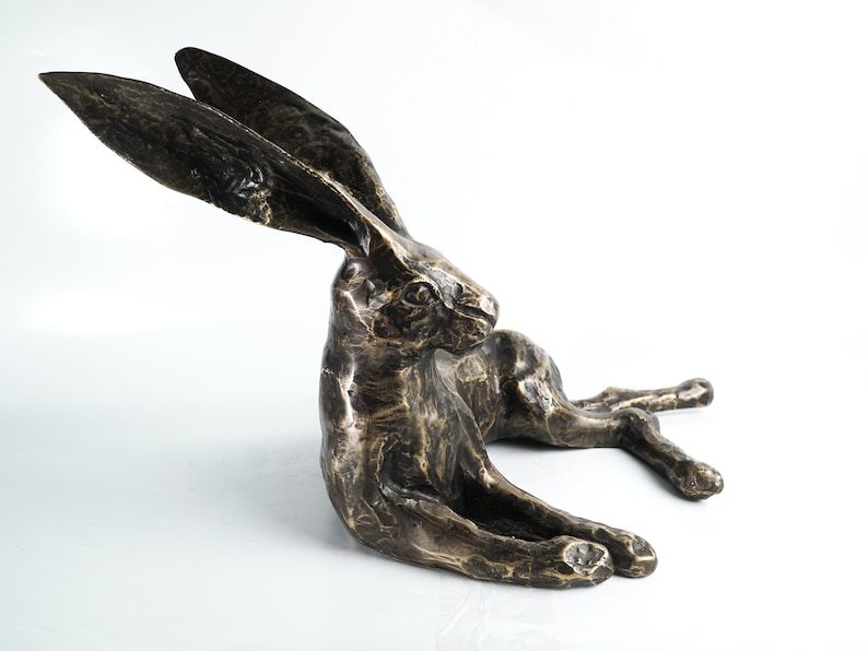 K&ouml;nnte beinhalten: Eine Bronzeskulptur eines liegenden Hasen mit seitlich gedrehtem Kopf. Der Hase hat lange Ohren und eine detaillierte, strukturierte Oberfl&auml;che.