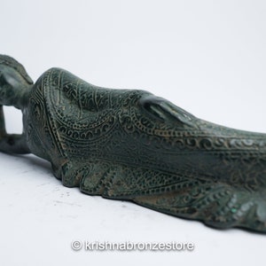May include: A green bronze statue of a reclining Buddha with intricate carvings. The statue is lying on its side with its head resting on its hand.