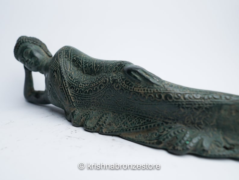 May include: A green bronze statue of a reclining Buddha with intricate carvings. The statue is lying on its side with its head resting on its hand.