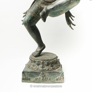 May include: A green bronze statue of a Hindu deity standing on a square base with intricate carvings. The statue is in a dancing pose with one leg raised.