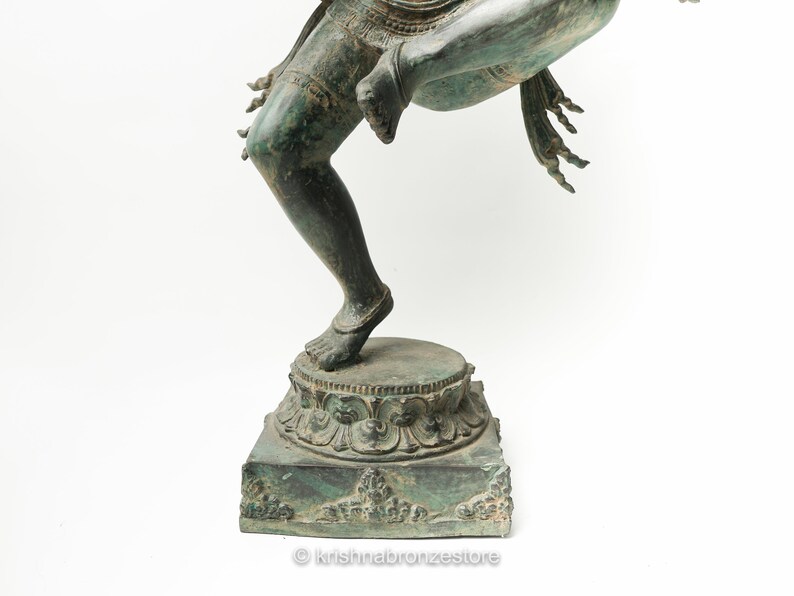 May include: A green bronze statue of a Hindu deity standing on a square base with intricate carvings. The statue is in a dancing pose with one leg raised.