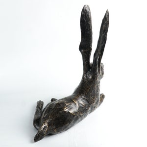 K&ouml;nnte beinhalten: Eine Bronzeskulptur eines liegenden Hasen mit erhobenen Ohren. Die Skulptur hat eine strukturierte Oberfl&auml;che und eine dunkelbraune Farbe.